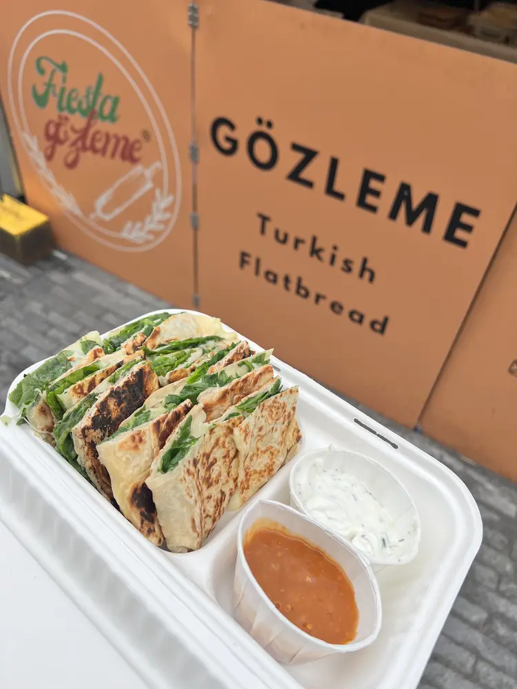 Sliced spinach and cheese gozleme in a takeaway box with hot sauce and garlic yoghurt, in front of the orange Fiesta Gozleme sign