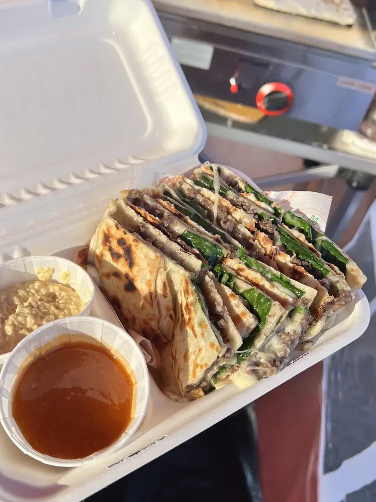A hand holding an open box of beef, spinach and cheese gozleme with hummus and hot sauce